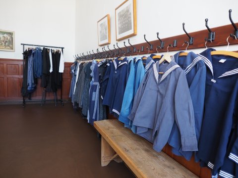 Alte Schuluniformen im Schulmuseum Bremen. Die Uniformen hängen aufgereiht an der Wand, unterteilt in Uniformen für Jungs und Uniformen für Mädchen